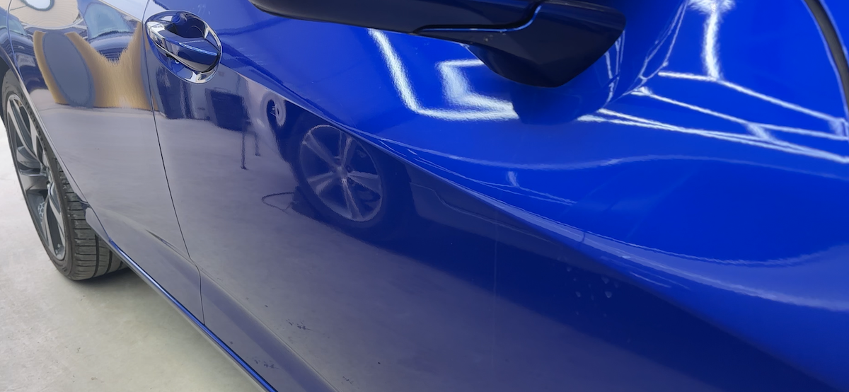 Before paintless dent repair showing distorted reflection lines on blue sedan panel in Aubrey Tx