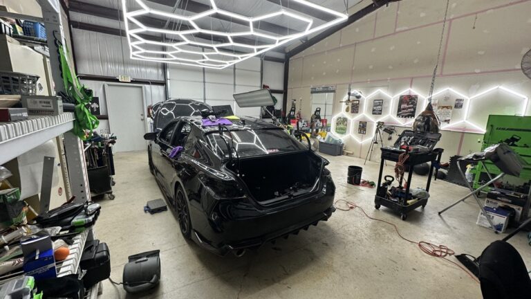 Black sedan with extensive hail damage undergoing Paintless Dent Repair inside the Divine Dents shop using specialized PDR lighting to evaluate and repair the vehicle roof.